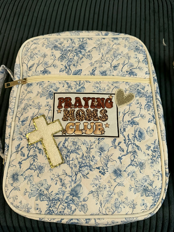 A blue and cream floral book cover with a chenille club patch on the front and a small cross charm on the zip.