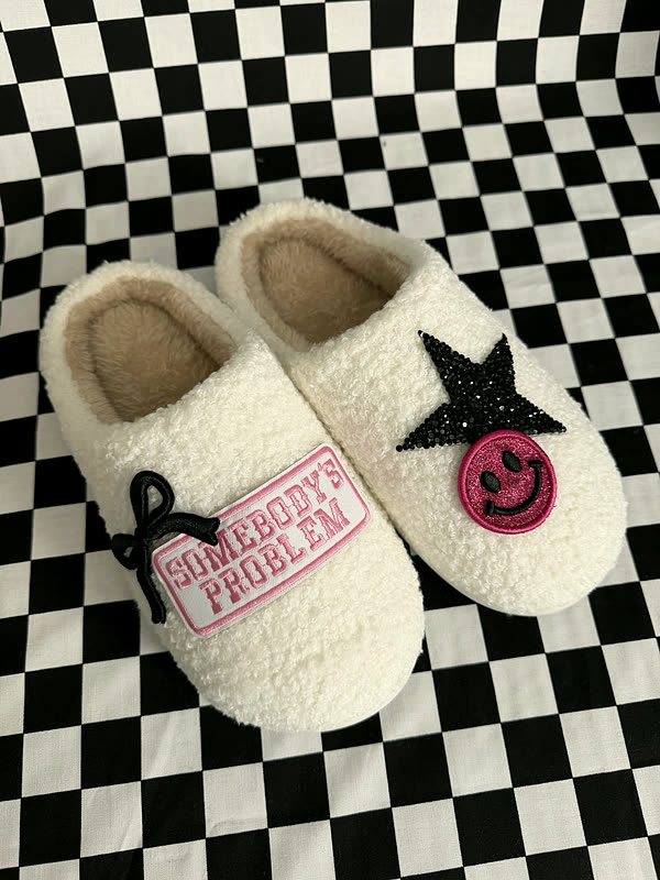 A pair of cream sherpa slippers on a checkered cloth, one with a black star, the other with a smiley and a word patch.