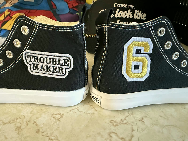 A pair of black canvas high-tops seen from behind, one with a word patch and the other with a chenille number.