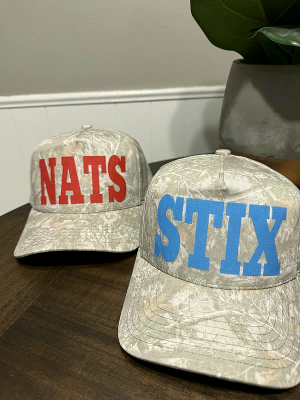 Two pale camo caps side by side, each with a different name in chenille letters across the front.