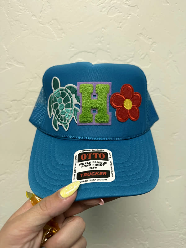 A bright blue cap with a chenille letter, a turtle patch and a red flower, held in one hand.