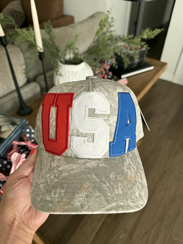 A pale camo cap with letter patches in stars and stripes across the front, held up in a styled room.