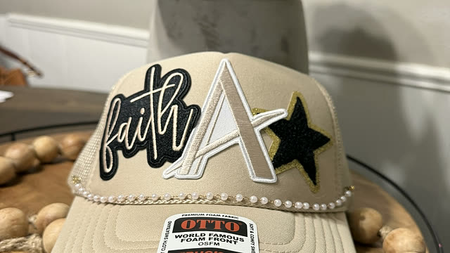 A tan trucker cap patched with a script “faith”, a monogram A and a black star, on a pearl brim trim